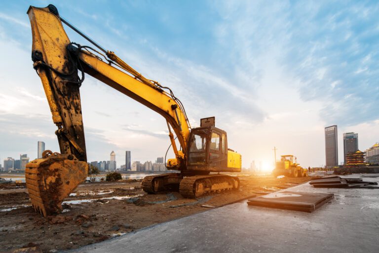 Excavator on construction site surrounded by high rise buildings with blue sky and sun setting