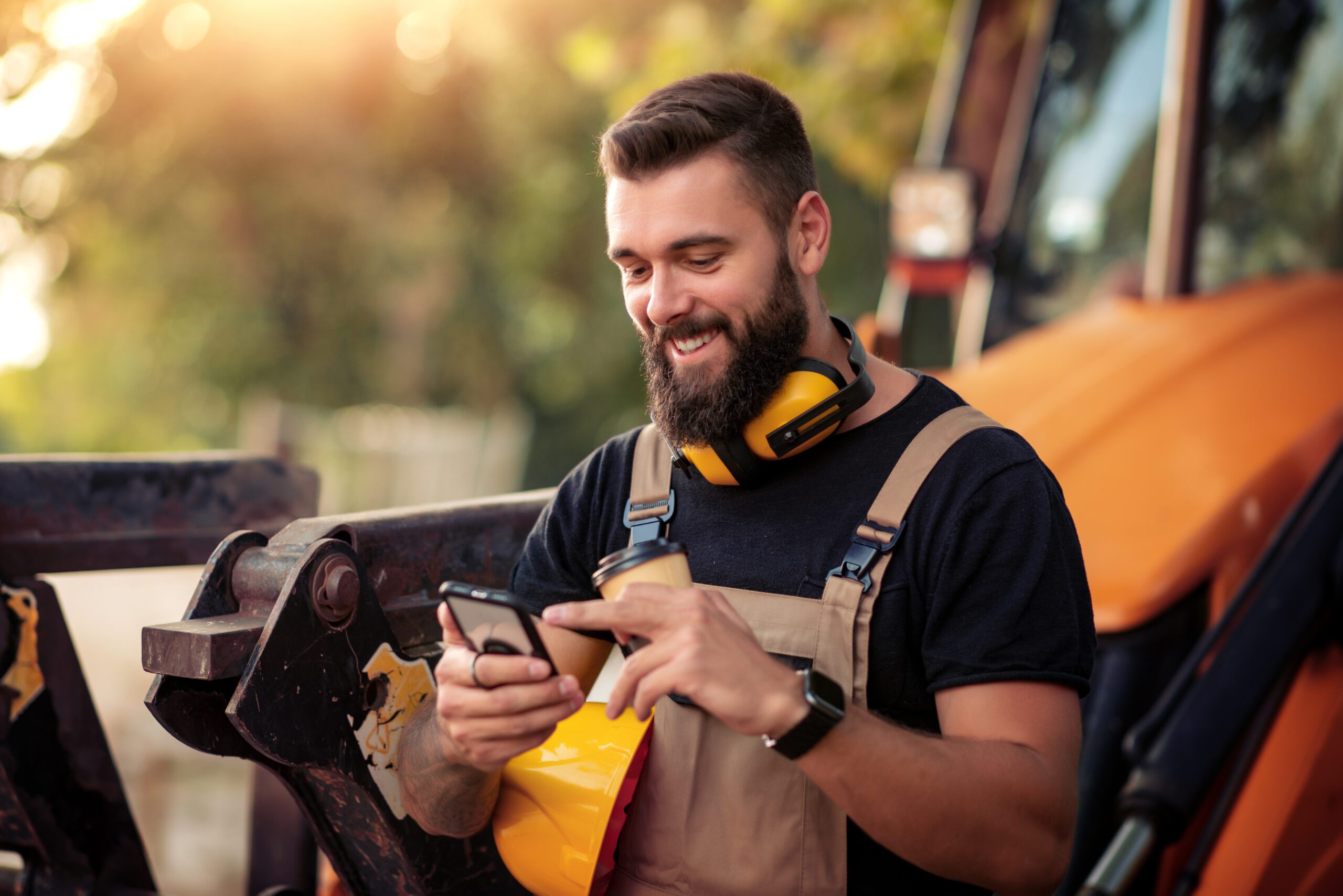Young male worker taking a break smiling at his mobile phone and leaning on heavy machinery