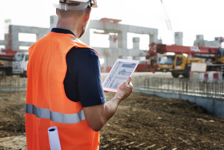 Construction contractor in orange hi-vis jacket looking at site plans on tablet with building site in the background